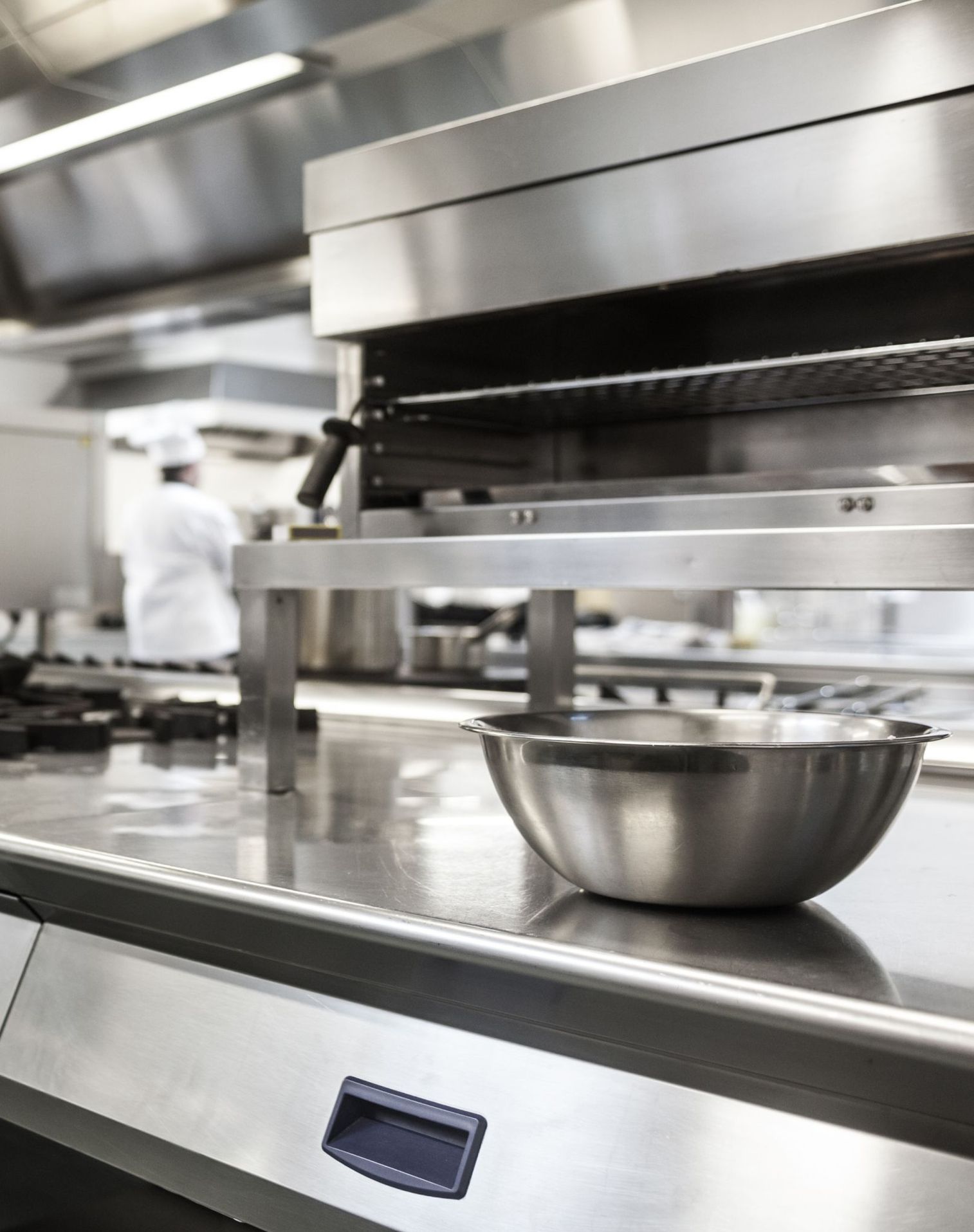 Edelstahlschüssel auf einer Edelstahlarbeitsfläche in einer professionellen Großküche mit verschwommenem Koch im Hintergrund