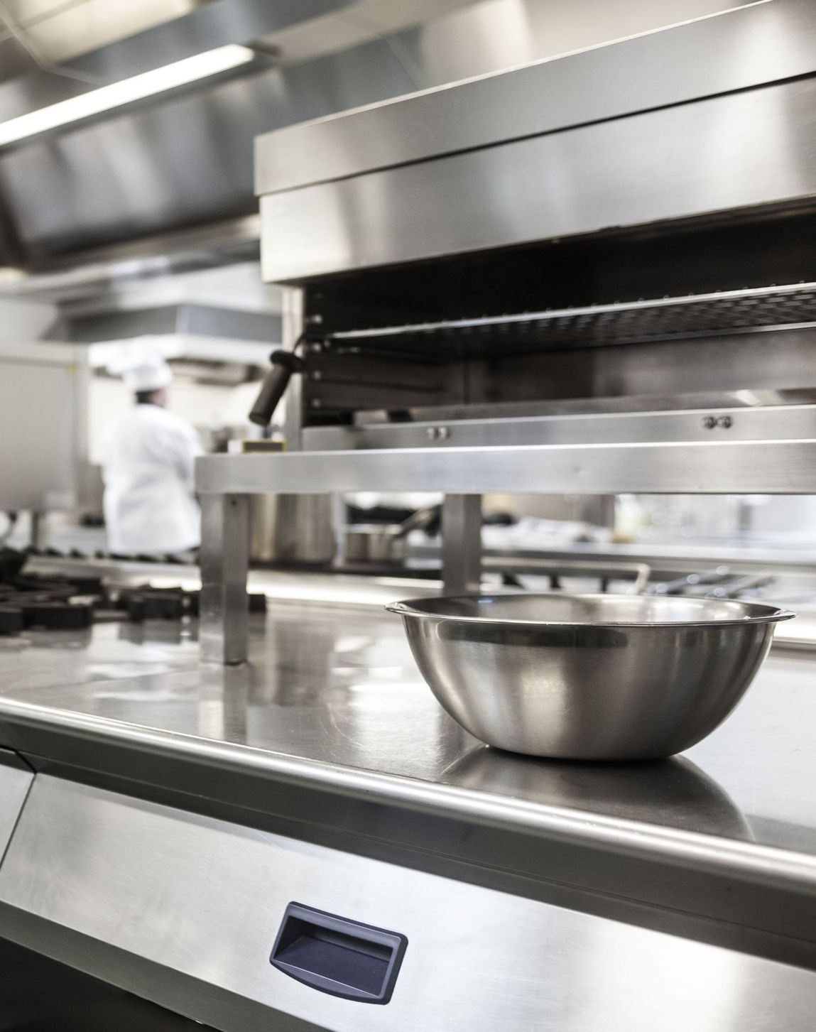 Edelstahlschüssel auf einer Edelstahlarbeitsfläche in einer professionellen Großküche mit verschwommenem Koch im Hintergrund Edelstahlschüssel auf einer Edelstahlarbeitsfläche in einer professionellen Großküche mit verschwommenem Koch im Hintergrund